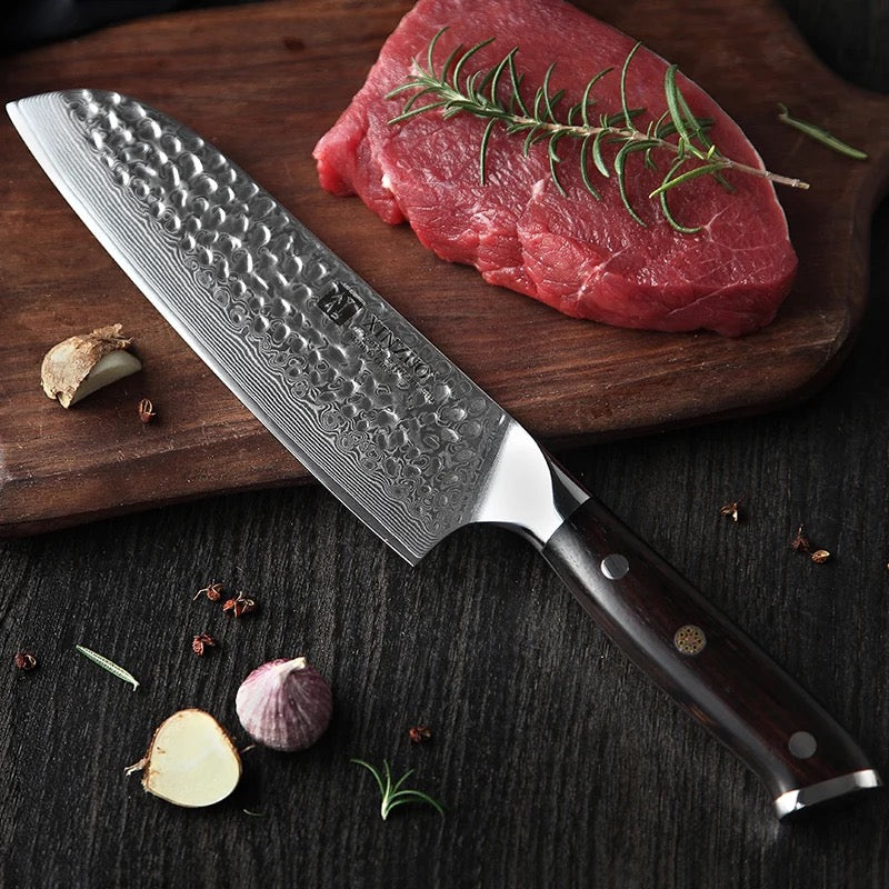 Cuchillo Xinzuo Santoku de 7 pulgadas en acero Damasco con mango oscuro junto a un corte de carne y romero.